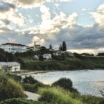 A view from a grassy hillside overlooking a coastal town with white buildings and red roofs nestled along a beach at sunset.
