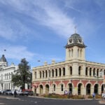 A street corner view of two grand, historic buildings with ornate classical architecture and prominent clock towers under a wide blue sky.