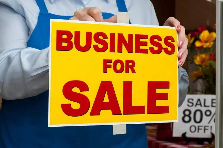 A person in a blue apron holding a large yellow sign with the bold red text "BUSINESS FOR SALE."