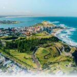 An aerial view of a sprawling coastal city nestled on a peninsula, bordered by blue ocean waters and sandy beaches under a bright, clear sky.