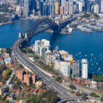 An aerial perspective of the Sydney Harbour Bridge connecting the city skyline to North Sydney over blue water.