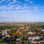 An aerial view of a town featuring a circular road layout and a long, straight avenue stretching toward the horizon under a wide, blue sky with scattered clouds.