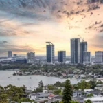 A wide skyline view of modern skyscrapers overlooking a body of water and a residential area during sunset