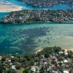 An aerial view of a coastal town featuring turquoise water and clear, shallow sandbars between lush, residential headlands.