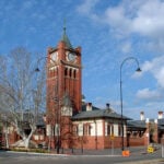 A historic red-brick building with a tall, prominent square clock tower under a clear blue sky with light clouds.