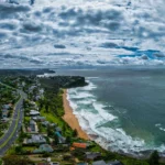 A high-angle view of a golden sand beach and crashing white waves along a coastal road lined with houses under a dramatic, cloudy sky.