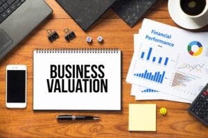An overhead shot of a wooden desk featuring a spiral-bound notebook with the bold black text "BUSINESS VALUATION".