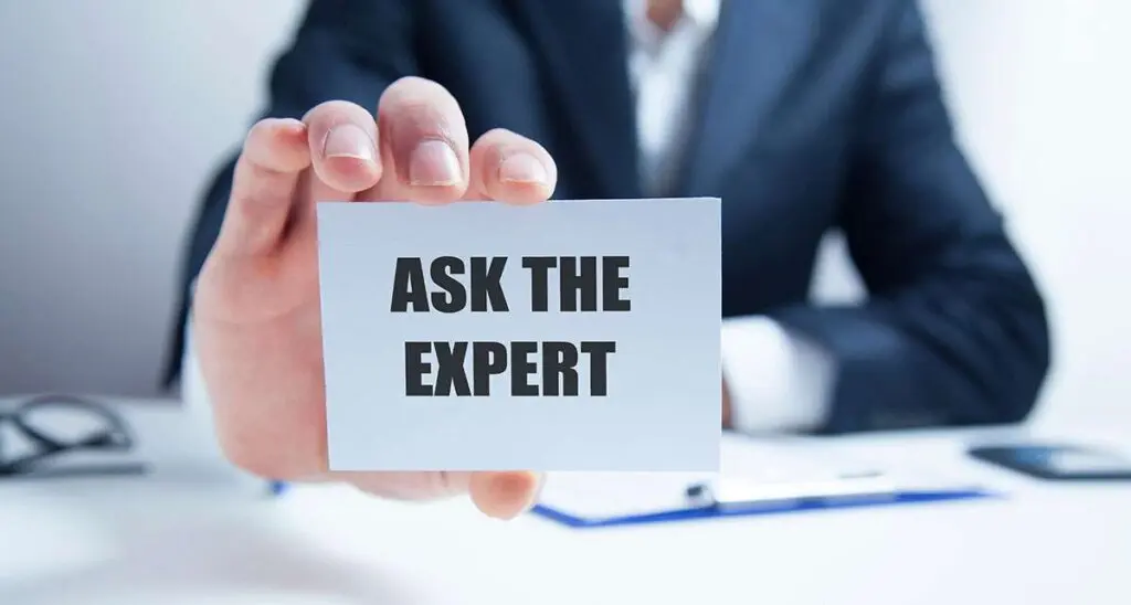 A person in a business suit holding a small white card towards the camera that reads "ASK THE EXPERT"