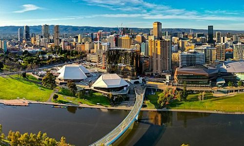 An aerial panorama of a city skyline nestled against a backdrop of rolling hills under a bright, clear sky.