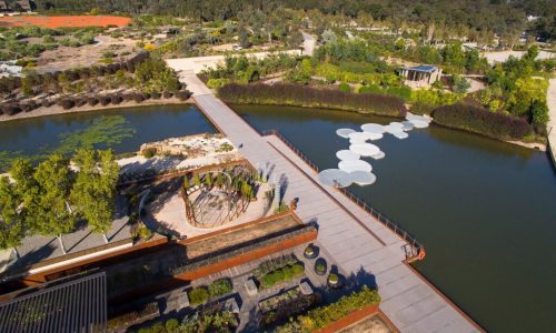 An aerial view of a botanical garden featuring a long pedestrian bridge crossing over a calm lake, with unique circular stepping stone platforms floating on the water.