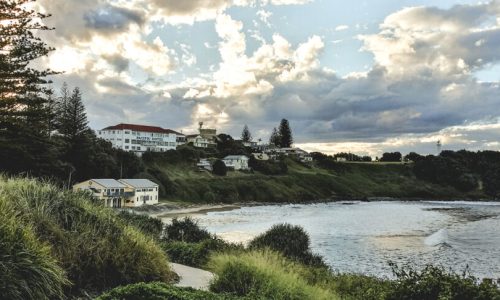 A view of a coastal bay at dusk featuring the white "Pacific Hotel" situated on a grassy hillside overlooking a sandy beach and rolling waves.