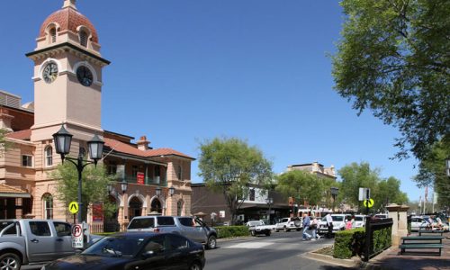 A street-level view of a sunny town centre featuring a grand pink historic building with a prominent domed clock tower on the left.