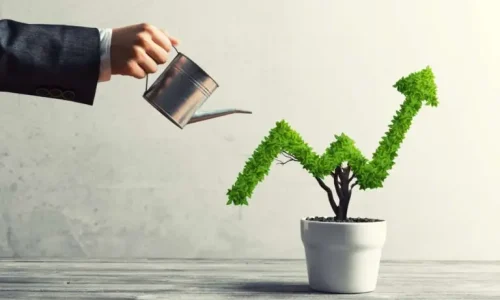 A conceptual image of a person in a business suit holding a small metal watering can, watering a plant growing in a white pot.
