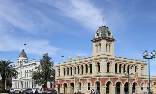 A street-level view of a grand, two-storey historic building with a prominent corner clock tower and arched colonnades on both levels.