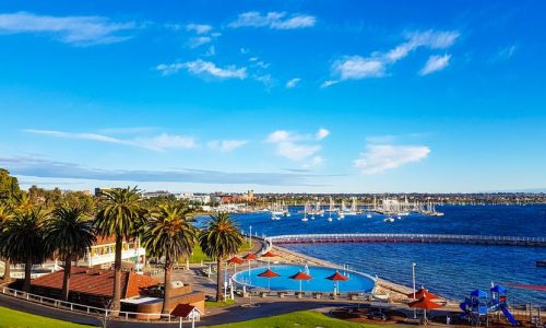 An elevated view of a coastal waterfront park featuring a circular blue swimming pool and a children’s playground with red sunshades.