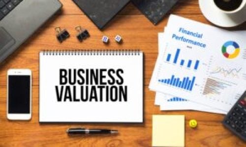 An overhead shot of a wooden desk featuring a spiral-bound notebook with the bold black text "BUSINESS VALUATION".