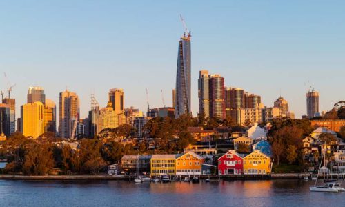 A scenic view across a blue harbor toward a city skyline at sunset, with a prominent tall skyscraper under construction in the center.