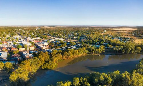 An aerial view of a small inland town situated alongside a calm river, with dense green trees lining the water's edge.