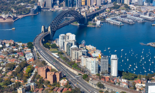 An aerial view of the Sydney Harbour Bridge spanning across a blue harbour, connecting a busy city skyline with a residential area.