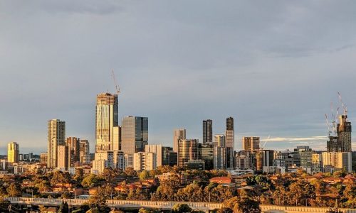 A panoramic view of a modern city skyline at sunset, featuring several high-rise glass buildings and a prominent skyscraper under construction with a crane at its peak.