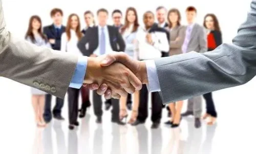 A close-up of two people in business suits shaking hands in the foreground, with a large, diverse group of blurred business professionals