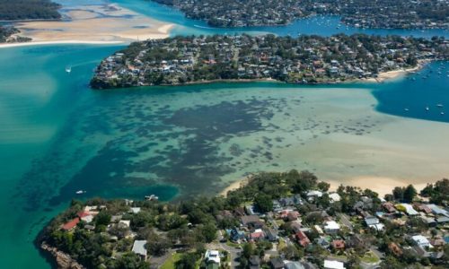 An aerial view of a coastal suburb featuring residential houses built on a peninsula and along the shoreline of a turquoise bay.