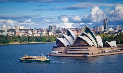 An elevated view of the iconic Sydney Opera House with its distinct white sail-like shells, situated on the edge of a blue harbor under a dramatic, cloudy sky.
