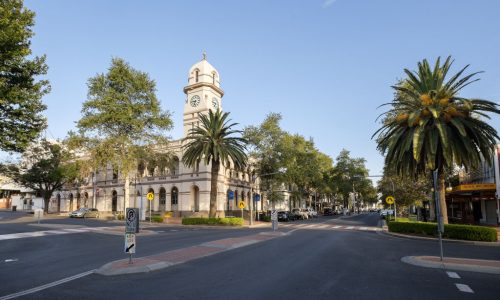 A street-level view of a grand white historic building with a multi-tiered clock tower and arched colonnades on both levels.