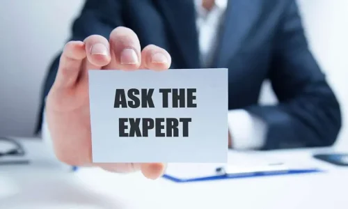A person in a business suit holding a small white card towards the camera that reads "ASK THE EXPERT"
