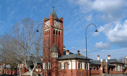 A street-level view of a grand, red brick historic building with a tall, central clock tower topped with a green spire.