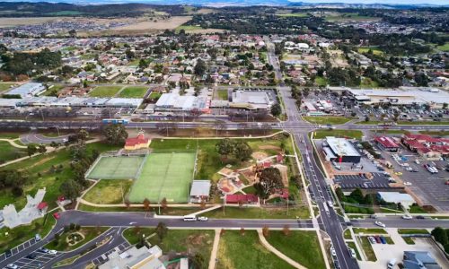 An aerial view of a sprawling suburban town, featuring a large green park with tennis courts and a playground in the foreground.