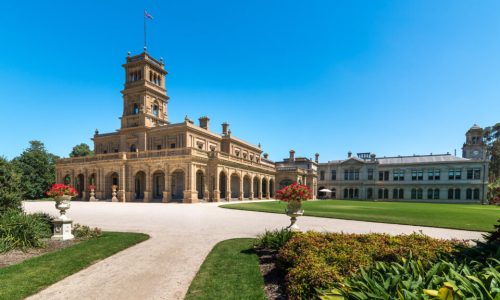 A ground-level view of a grand, sand-coloured historic mansion featuring a tall central tower with a flag