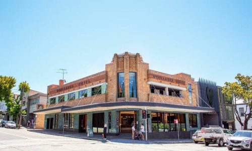 A ground-level view of the two-story brick Woollahra Hotel, featuring an Art Deco facade with a curved corner and blue tiling on the lower level.