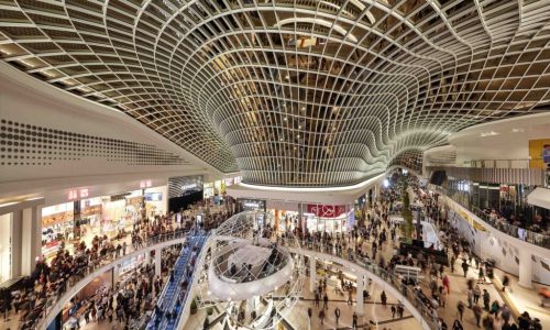 An interior view of a vast, modern multi-level shopping mall featuring a striking, wave-like glass and steel lattice ceiling.