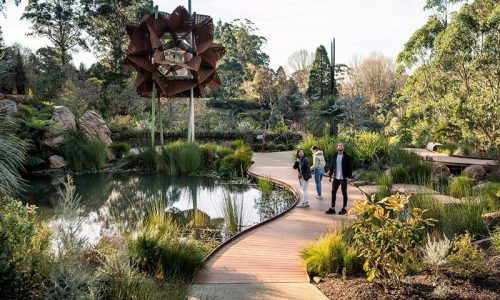 A view of a winding wooden boardwalk through a lush botanical garden, featuring three people walking past a small pond and a large, abstract metal sculpture.