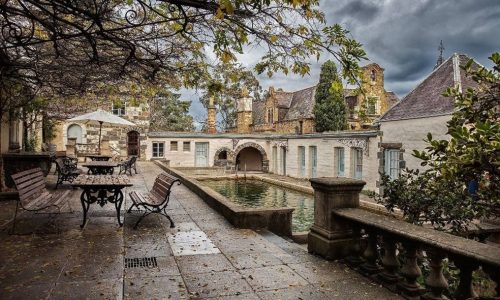 A view of a rustic stone patio and courtyard featuring a narrow swimming pool, iron benches, and several historic, cottage-style buildings under a dark, cloudy sky.