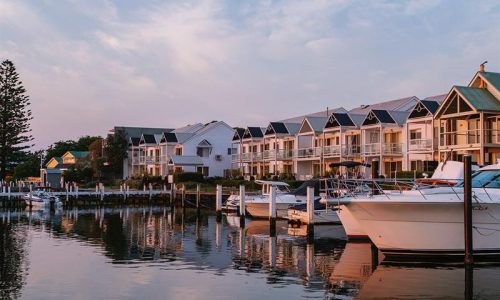 A row of modern, white two-storey townhouses with gabled roofs overlooks a calm marina at sunset.